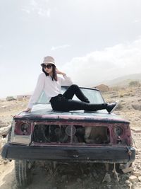 Fashionable woman sitting on abandoned car against sky