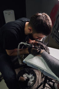Concentrated male tattooist in gloves making tattoo on hand of client while using professional tattoo machine in modern tattoo studio