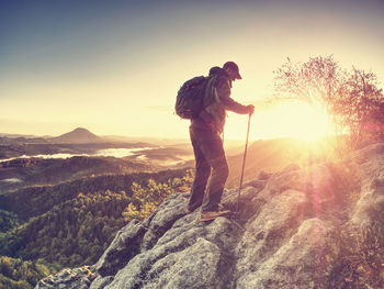 Man on rocky cliff climbing up with trekking poles and backpack, sunset behind man.