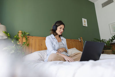 Smiling pregnant businesswoman wearing wireless headphones using laptop on bed in bedroom