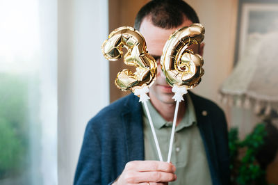 Man in a blue jacket holds in his hands little foiled gold balloons of the number 36 on sticks 