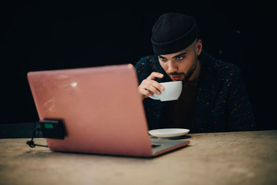 Confident young male hacker drinking coffee while looking at laptop on desk in office