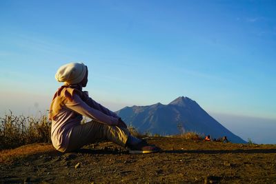 Side view of woman sitting on mountain against sky at sunset