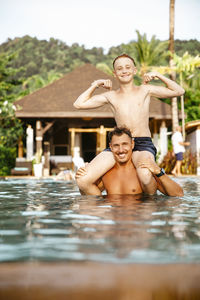 Portrait of father carrying son on shoulders in swimming pool during vacation