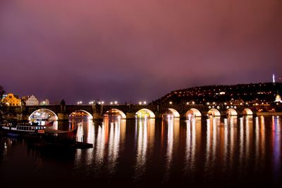 Illuminated bridge over river by buildings against sky at night