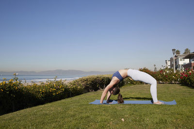 Full length of woman bending over backwards on exercise mat against clear sky