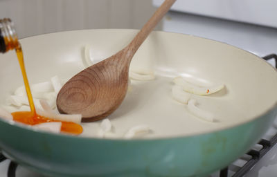 Orange cooking oil pouring over sliced white onion in a frying pan with a wooden spoon