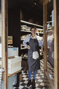 Portrait of smiling young female owner standing by door of delicatessen shop