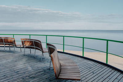 Empty deck chairs by sea against sky