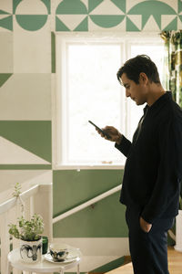 Side view of man with hand in pocket using smart phone while standing at home