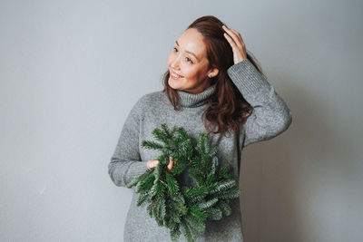 Adult woman in warm grey dress with diy fir christmas wreath in hand on grey background