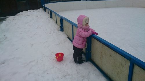 Side view of girl standing on snow covered road by fence during winter