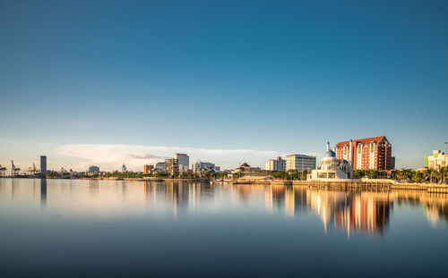 Buildings by river against clear blue sky