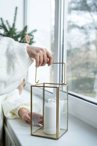 Woman in warm white winter sweater sitting on the window at home at christmas eve holding cup 