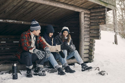 Friends sharing drink while sitting at log cabin during winter