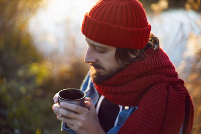 Man in a red knitted scarf and hat stands by the lake in autumn and drinks tea mug thermos