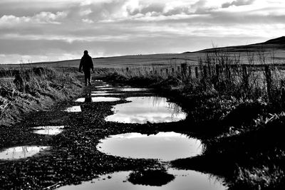 Rear view of silhouette man walking by puddle on pathway against sky