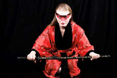 Portrait of young female model holding samurai sword while kneeling against black curtain