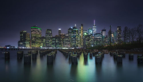 Illuminated modern buildings by river against sky at night