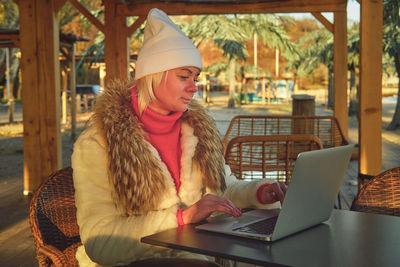 A woman in a sweater and a fur coat is sitting on the terrace of a cafe with a laptop.