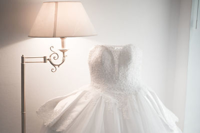 Wedding dress by illuminated lamp against white wall at home