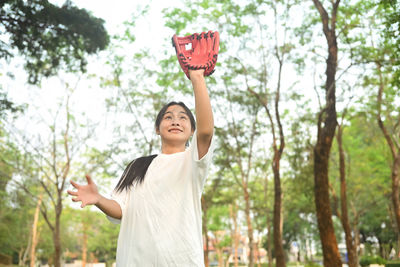 Portrait of young woman with arms raised standing against trees