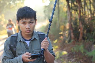 Portrait of young man holding mobile phone with monopod while hiking in forest