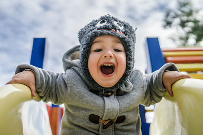 Low angle portrait of happy baby boy screaming while sliding against sky at playground