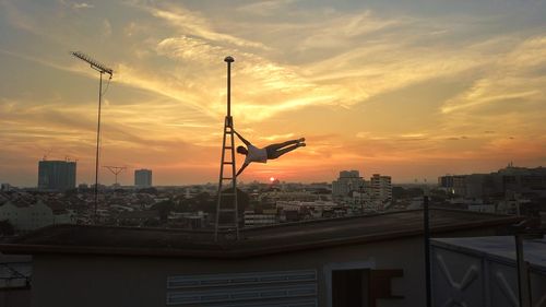 Man performing stunt on terrace at cityscape against sky during sunset