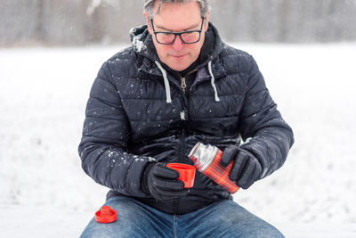 Man holding coffee in thermos while sitting outdoors during winter