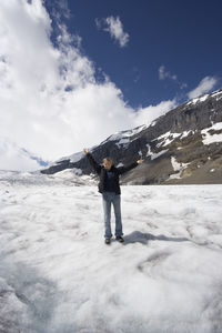Happy young woman with arms raised standing on snow covered mountain against sky