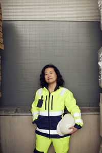 Portrait of confident female worker in reflective clothing standing against wall in factory