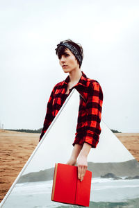 Serious pensive young female in casual red and black checkered shirt standing on sandy beach with red book in hand and large mirror with sea and rocks reflection