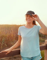 Smiling teenage girl standing near the field wearing light blue t-shirt, baseball cap