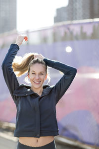 Portrait of confident athlete tying hair while standing by fence during sunny day