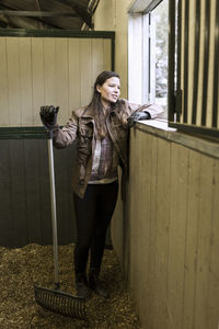 Full length of young woman with pitchfork looking through window of horse stable