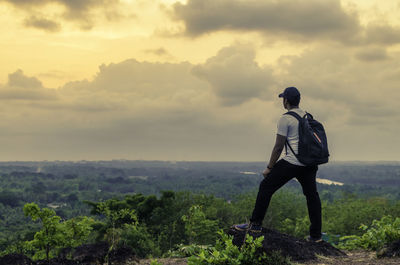 Rear view of man standing on cliff against cloudy sky during sunset