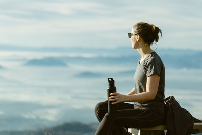 Young woman looking at view while sitting on bench against sky