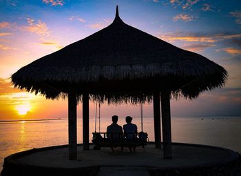 Silhouette couple sitting under gazebo at beach against sky during sunset