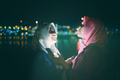 Smiling friends wearing hijabs using mobile phones while standing against sky at night