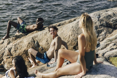 Smiling shirtless man talking with female friend sitting on rock near lake