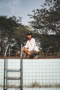 Young woman crouching at abandoned swimming pool against sky