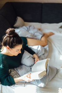High angle view of young woman reading book while lying on bed at home