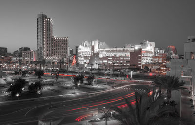 Light trails on city street by buildings against sky at night