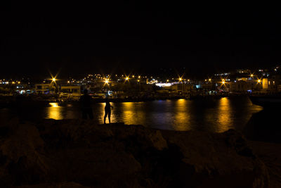 Illuminated city by sea against sky at night