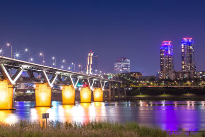 Illuminated bridge over river by buildings against sky at night