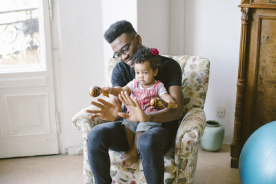 Baby girl holding maracas while sitting with father on armchair at home