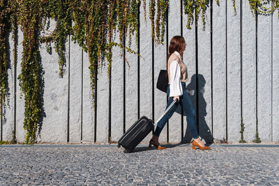 Side view of positive young redhead woman with a black suitcase walking in the city