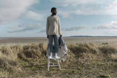 Young man with blanket standing on stool while looking away against cloudy sky