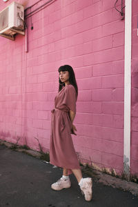 Side view of stylish young ethnic female with long dark hair in trendy dress standing against pink wall on street and looking away thoughtfully